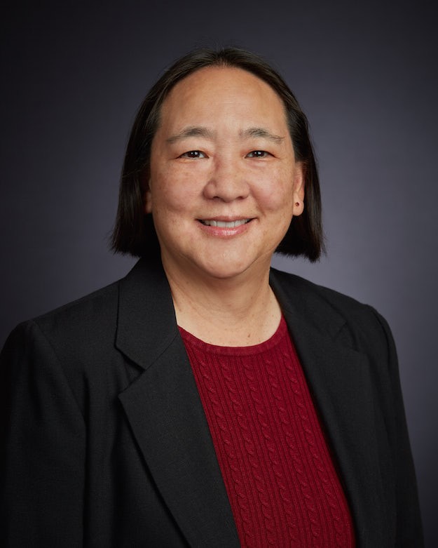 Ann West has shoulder-length dark hair and is smiling. They are wearing a black blazer over a red sweater. The background is dark gray, giving a professional tone.